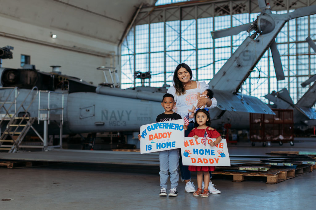 Woman, 2 kids and a baby holding "Welcome Home" signs in front of a Navy Helicopter for a Military Homecoming