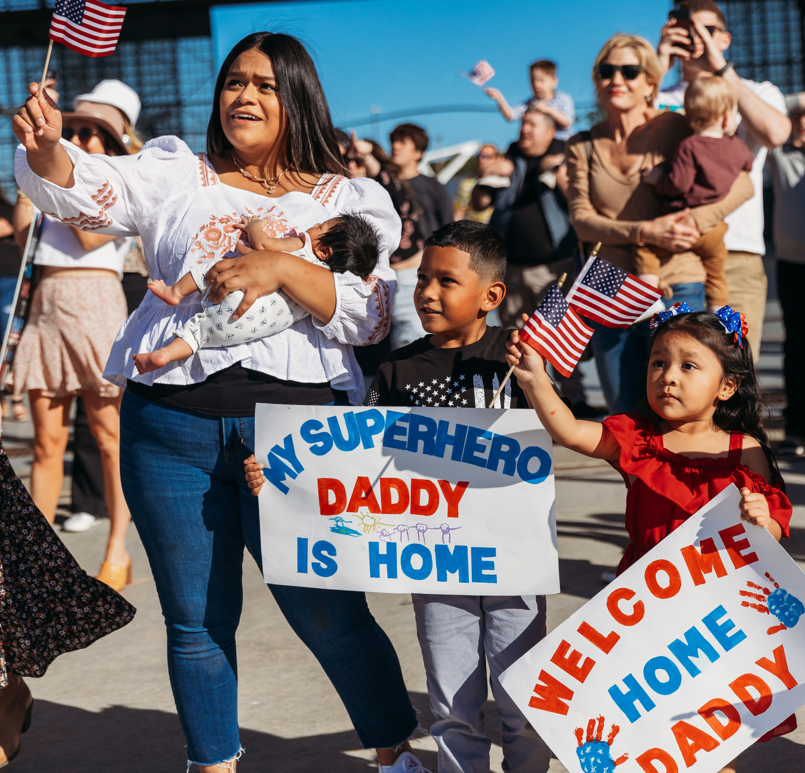 Woman and kids hold American Flags and Welcome Home signs at a military homecoming
